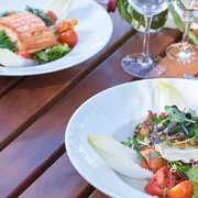 Restaurant Mediterraneo KOCHKULTUR Nahaufnahme von einem Holztisch mit leeren Weingläsern und zwei Tellern. Auf dem einen Teller ist gekochter Lachs mit Salat und auf dem anderen ein Salat mit Ziegenkäse und Chicoree.