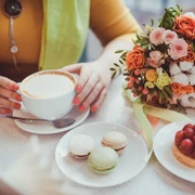 Runder Café-Tisch mit einem bunten Blumenstrauß, einem Erdbeertörtchen, drei Macarons und einem Cappuccino, den eine Frau mit der Hand festhält.