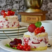 Nahaufnahme von einem Teller, auf dem ein rundes Stück Kuchen mit Erdbeeren und Johannisbeeren steht. Im Hintergrund stehen gestapelte Teller, wobei auf dem oberen auch ein solches Kuchenstück liegt.