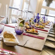 Langer gedeckter Tisch mit Besteck, Servietten, Gläsern und Tellern mit Brot sowie Käse und Trauben. Am Tisch stehen mehrere Stühle.