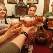 Junge Menschen stoßen lachend mit vollen Biergläsern an einem Holztisch in einer rustikalen Umgebung an.