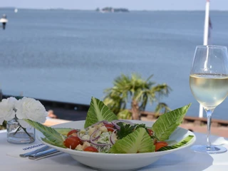 Ein frischer Salat mit Tomaten und Gurken auf einem Tisch mit Seeblick und einem Glas Weißwein.