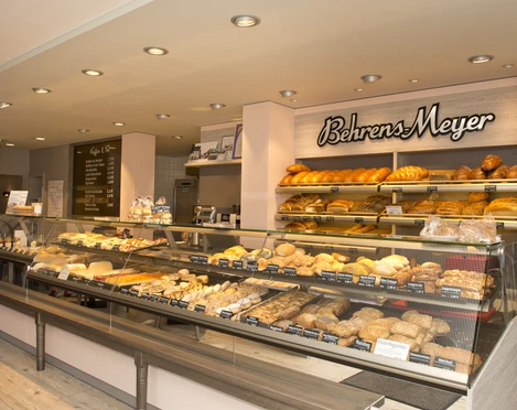 Innenansicht Cafe Behrens-Meyer Die Innenansicht des Cafés Behrens-Meyer zeigt eine einladende Theke, voll bestückt mit frischen Backwaren. An der Wand dahinter sind dekorativ das Logo des Cafés und verschiedene köstlich aussehende Brotsorten arrangiert.