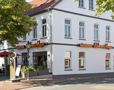 Aussenansicht Cafe Behrens-Meyer Das Bild zeigt die Außenansicht des Cafés Behrens-Meyer, ein charmantes weißes Gebäude mit großen Fenstern, umgeben von Bäumen und einer einladenden Terrasse mit Sonnenschirmen. Leuchtende Schilder weisen auf die Bäckerei hin.