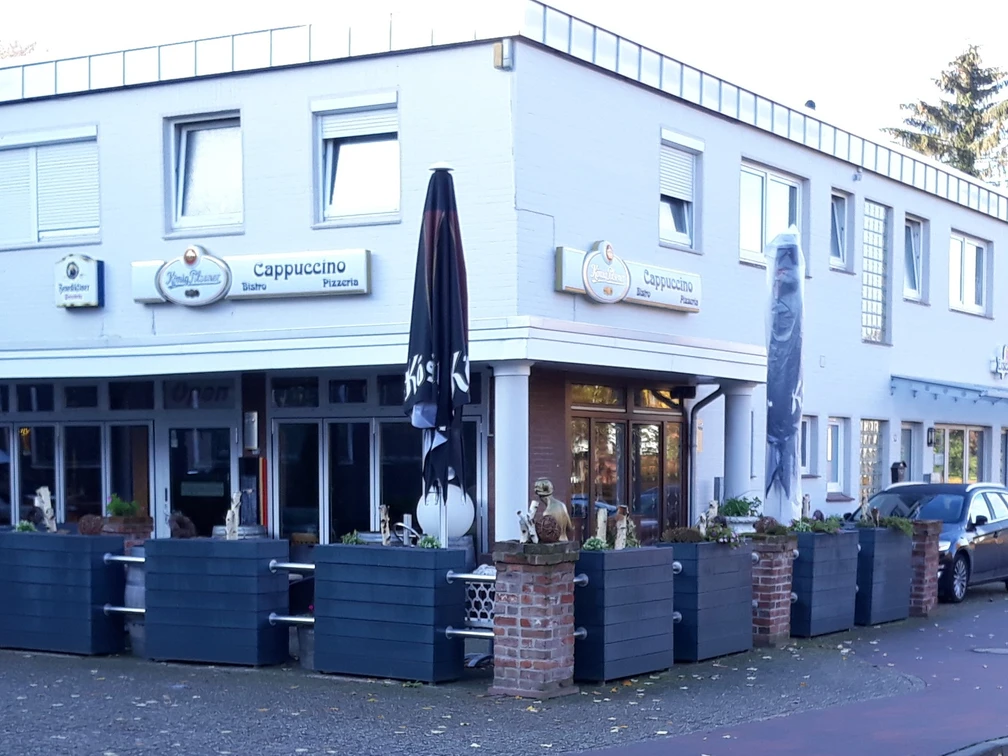 Bistro Cappuccino Das Bild zeigt das Bistro Cappuccino, einen zweistöckigen, weiß verputzten Bau mit markanten schwarzen Sonnenschirmen und modernen Außensitzbereichen, die mit Pflanzkästen geschmückt sind. Im Hintergrund parkt ein Auto. Die Schilder über den Fenstern tragen den Bistro-Namen.