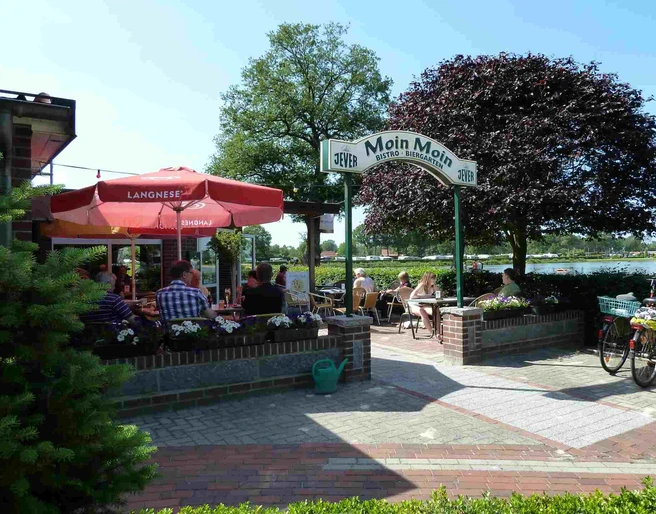 Bistro Moin Moin Vor dem "Bistro Moin Moin" sitzen Gäste unter einem roten Sonnenschirm. Ein einladender Eingang mit Blumen führt in das gemütliche Café. Fahrräder sind im Vordergrund abgestellt, ein schöner Tag am Wasser.