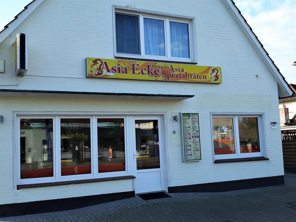 Asia Ecke Das Bild zeigt ein kleines, weißes Gebäude mit einem Giebel, das ein asiatisches Restaurant beherbergt. Über dem Eingang hängt ein Schild mit der Aufschrift „Asia Ecke – Asia Spezialitäten“, flankiert von roten Drachenmotiven. Große Fenster in der Front verheißen ein helles Inneres, während das Kopfsteinpflaster den rustikalen Charme des Ortes unterstreicht.