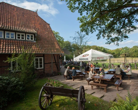 gasthaus-zum-heidemuseum Das Bild zeigt ein traditionelles, reetgedecktes Gasthaus aus rotem Backstein. Vor dem Gebäude sitzen Gäste an Holztischen im Freien unter einem weißen Sonnenschirm. Links im Vordergrund befindet sich ein alter Holzkarren auf einer Rasenfläche. Im Hintergrund erstreckt sich eine Wiese.