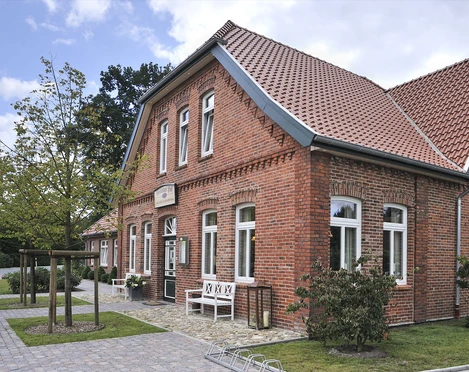 Gristeder Hof Das Bild zeigt ein hübsches, großes Gebäude aus rotem Backstein mit modernen weißen Fensterrahmen. Es ist der Gristeder Hof, ein traditionelles, zweigeschossiges Haus mit einem steilen Ziegeldach. Vor dem Gebäude läuft eine gepflasterte Fläche entlang, auf der sich Fahrradbügel und eine Bank befinden. Ein kleiner Baum steht am linken Rand. Im Hintergrund ist Himmel mit vereinzelten Wolken zu sehen.
