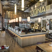Barhocker stehen an einer großen Theke, in der sich Gläser, Tassen, eine Kaffeemaschine und ein Regal mit weiteren Gläsern und Flaschen befinden. Über der Theke hängt die Leuchtschrift "CUCINA" und davor mehrere Tischgruppen. Von der Decke hängen Lampen herab.
