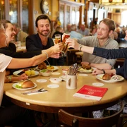 Früh am Dom Eine Gruppe von Menschen sitz an einem runden Tisch, stoßen mit Kölsch an und haben kleine kölsche Speisen vor sich stehen.A group of people are sitting around a round table, clinking glasses of Kölsch beer and having small Cologne dishes in front of them.