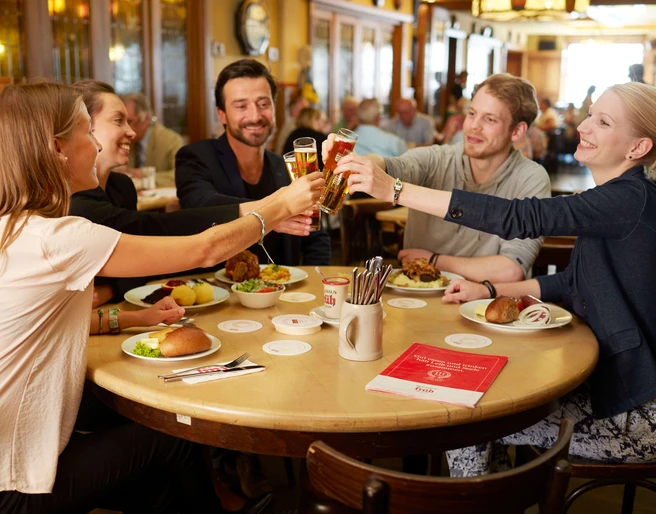 Früh am Dom Eine Gruppe von Menschen sitz an einem runden Tisch, stoßen mit Kölsch an und haben kleine kölsche Speisen vor sich stehen.A group of people are sitting around a round table, clinking glasses of Kölsch beer and having small Cologne dishes in front of them.