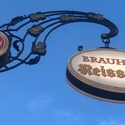 Reissdorf Zwei kunstvoll verzierte Brauhaus-Schilder, "Reissdorf Kölsch" und "Brauhaus Reissdorf", vor blauem Himmel.Two artistically decorated brewery signs, "Reissdorf Kölsch" and "Brauhaus Reissdorf", against a blue sky.