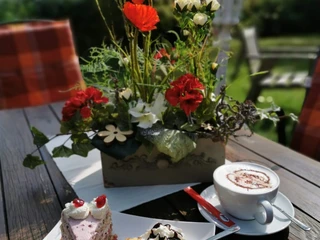 Floristik unterm Storchennest Blumenarrangement mit roten Mohnblumen, daneben Tortenstücke und eine Tasse Cappuccino auf Holztisch.