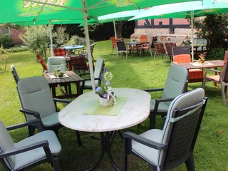 Floristik unterm Storchennest Ein Gartenbereich mit runden Tischen und Stühlen unter grünen Sonnenschirmen, umgeben von Pflanzen.