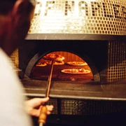 Ein Mann schiebt eine Pizza auf einer flachen Schaufel in einen großen Pizzaofen, in dem sich bereits weitere Pizzen befinden.