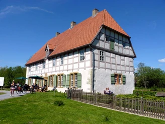 Fachwerkhaus mit rotem Ziegeldach und grünen Fensterläden, umgeben von Rasen und Besuchern im Freien.