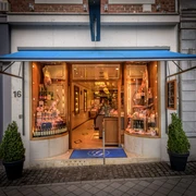 fleischerei-gerrards Der Eingang zur Fleischerei besteht aus Schaufenstern, hinter denen Flaschen und Wurstwaren ausgelegt sind, und einer Tür mit Teppich in der Mitte. Über dem Eingang ist eine Markise ausgefahren. Innen befindet sich die Fleischtheke sowie mehrere Wandregale.