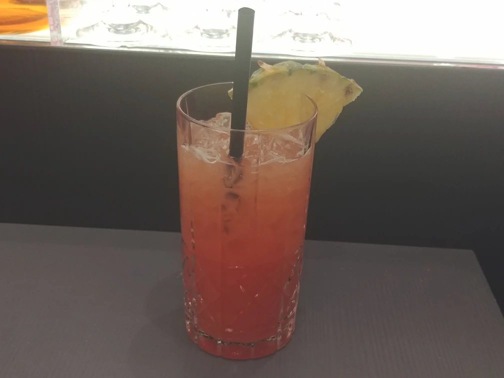 Ein erfrischender roter Cocktail in einem hohen Glas mit Ananasscheibe und Strohhalm darauf.