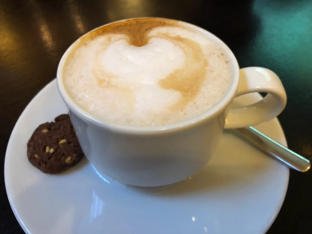 Cappuccino mit reichhaltigem Milchschaum und Keks auf einer weißen Untertasse, leicht von oben betrachtet.