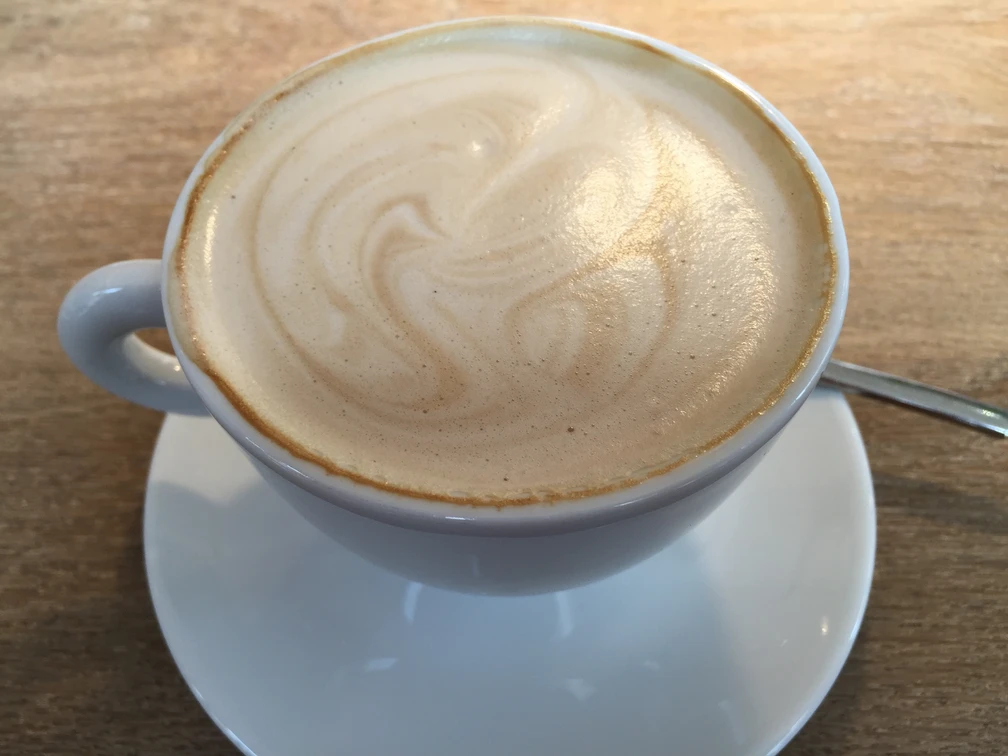 Kaffeetasse Kaffeetasse mit schaumiger Milchoberfläche auf Holztisch, begleitet von einem Teelöffel.