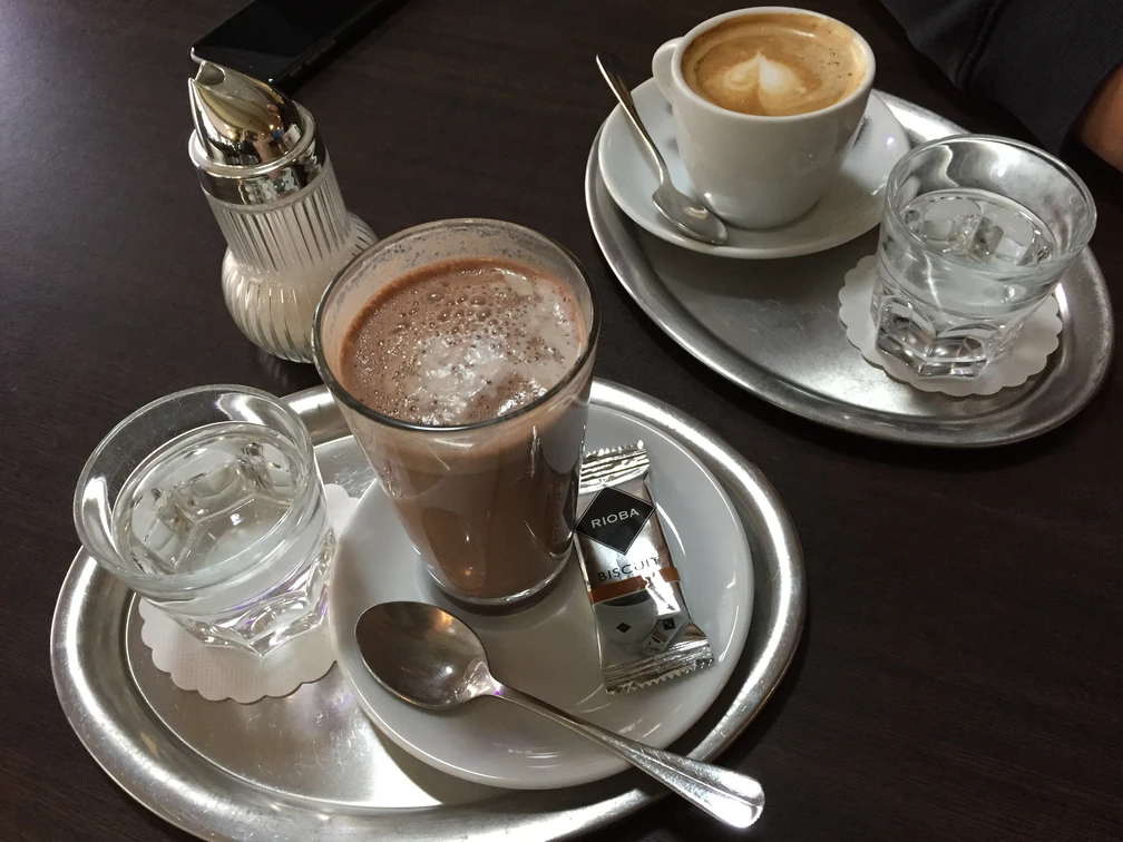 Ein Tablett auf dunklem Tisch mit einem Cappuccino, einem Glas Wasser und einem Schokoladengetränk.