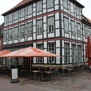Cup & Cino Fachwerkhaus mit Café-Terrasse, Stühle und Tische im Freien, bei blauem Himmel und roten Sonnenschirmen.