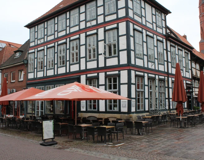 Cup & Cino Fachwerkhaus mit Café-Terrasse, Stühle und Tische im Freien, bei blauem Himmel und roten Sonnenschirmen.