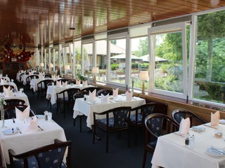 Großes Restaurant mit gedeckten Tischen, Panoramafront und Blick auf grüne Landschaft draußen.