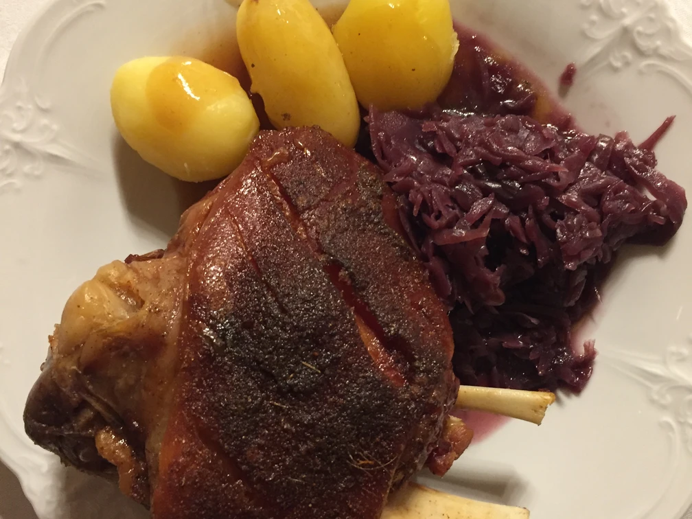 Haxe Gegrillte Schweinshaxe, serviert mit drei Salzkartoffeln und einer Portion Rotkohl auf einem weißen Teller.