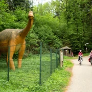 Dinosaurier bei den Dinosaurierspuren bei Barkhausen Zwei Radfahrer auf einem Waldweg, neben einer großen Dinosaurier-Statue im Grünen.