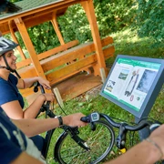 Hörstation auf der Ackerschnacker Tour Zwei Radfahrer betrachten eine Infotafel neben einem überdachten Holzunterstand im Grünen.