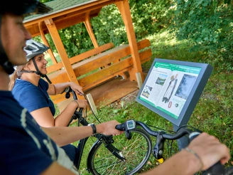 Hörstation auf der Ackerschnacker Tour Zwei Radfahrer betrachten eine Infotafel neben einem überdachten Holzunterstand im Grünen.
