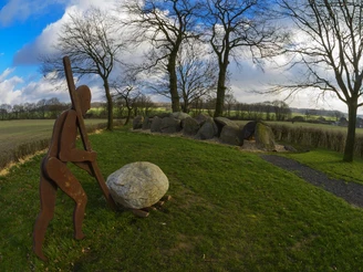 Radeln auf der Radroute der Megalithkultur Metallskulptur eines Menschen, der einen großen Stein inmitten einer grünen Wiese rollt.