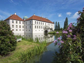 Schloss Hünnfeld im Osnabrücker Land Ein weißes Wasserschloss mit rotem Dach spiegelt sich im Wasser, umgeben von blühenden Sträuchern.