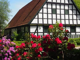 Radeln auf der BE 1 - Kleine Schlössertour Fachwerkhaus mit rotem Dach, umgeben von blühenden Rhododendren und Bäumen im Hintergrund.