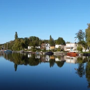Grachten in Großenheidorn Gracht mit ruhigem Wasser, umgeben von Bäumen und Häusern im Grünen, unter klarem, blauem Himmel.