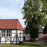 Eine kleine Fachwerkkirche mit roten Ziegeldach neben einem grünen Baum im Dorfzentrum.