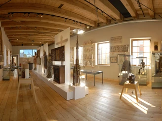 Museum Wiedenbrücker Schule Ein heller Museumsraum mit Holzdecke, Exponaten in Vitrinen und Skulpturen auf einem Podest.