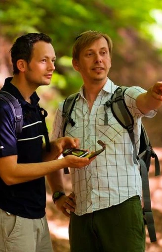 Unterwegs auf dem GPS-Erlebnispfad "Erlesene Natur - Waldwelten" Zwei Männer wandern auf einem Waldpfad unter grünen Blättern, einer zeigt lächelnd in die Ferne.