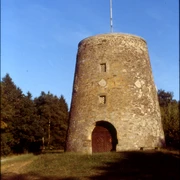 Kumsttonne_Herbstansicht Eine alte, steinerne Windmühle steht vor einem Hintergrund aus herbstlichen Bäumen unter klarem Himmel.