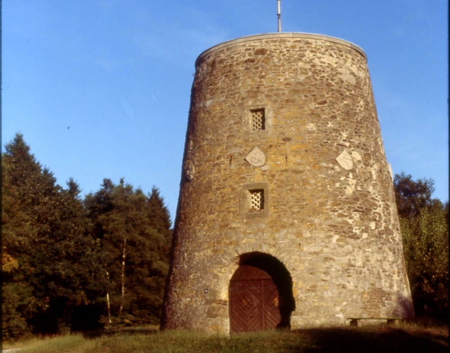 Kumsttonne_Herbstansicht Eine alte, steinerne Windmühle steht vor einem Hintergrund aus herbstlichen Bäumen unter klarem Himmel.