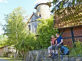 Levedags Mühle: Rast Zwei Personen sitzen auf einer Steinmauer vor einer alten Mühle, umgeben von grüner Vegetation.