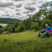 Wanderpaar bei der Rast  Ein Wanderpaar sitzt auf einer Wiese mit Rucksäcken, umgeben von hügeliger Landschaft und Wolkenhimmel.