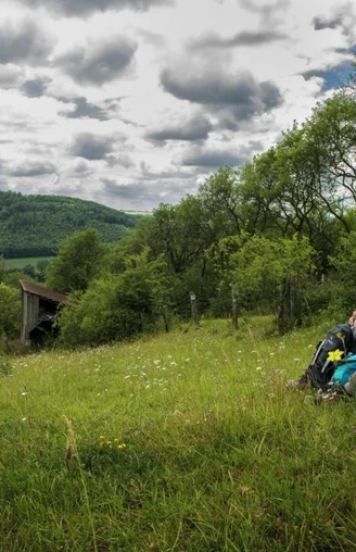 Wanderpaar bei der Rast  Ein Wanderpaar sitzt auf einer Wiese mit Rucksäcken, umgeben von hügeliger Landschaft und Wolkenhimmel.