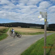 Ein Radweg in ländlicher Landschaft; Wegweiser; zwei Fahrräder und Personen auf einem asphaltierten Weg.