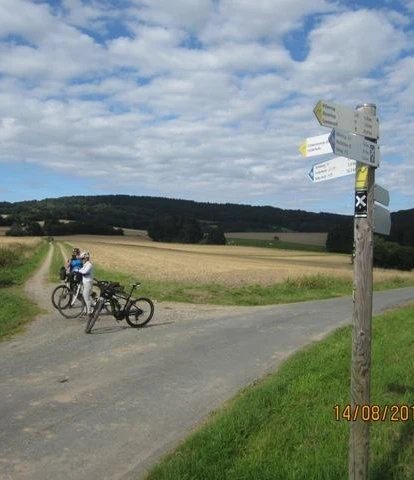 Ein Radweg in ländlicher Landschaft; Wegweiser; zwei Fahrräder und Personen auf einem asphaltierten Weg.