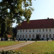 Historischer Gutshof mit rotem Dach, flankiert von Fachwerkgebäuden, umgeben von grüner Natur.