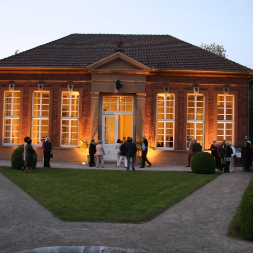 Orangerie Rotes Backsteingebäude mit großer Fensterfront, abends beleuchtet, Besucher im Vordergrund.