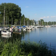 Segelboote liegen vertäut an einem Steg eines ruhigen Sees, im Hintergrund sind Bäume sichtbar.